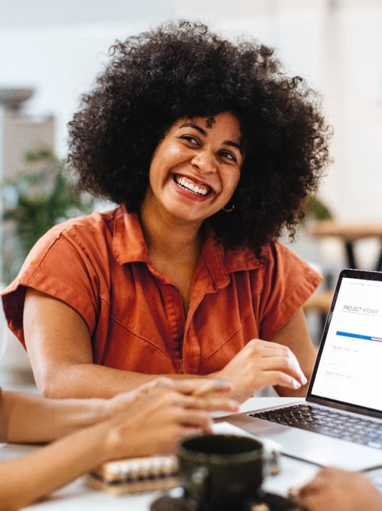 Smiling woman in a rust-colored shirt sitting at a table with a laptop, engaged in a collaborative meeting.